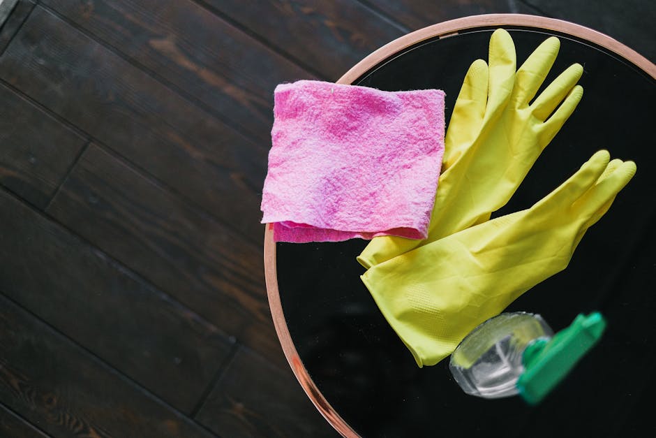 A person wearing blue and yellow rubber gloves is spray-cleaning and wiping down the surface of a white gas stove with black burners in a domestic kitchen. The stove appears to be in good condition, w