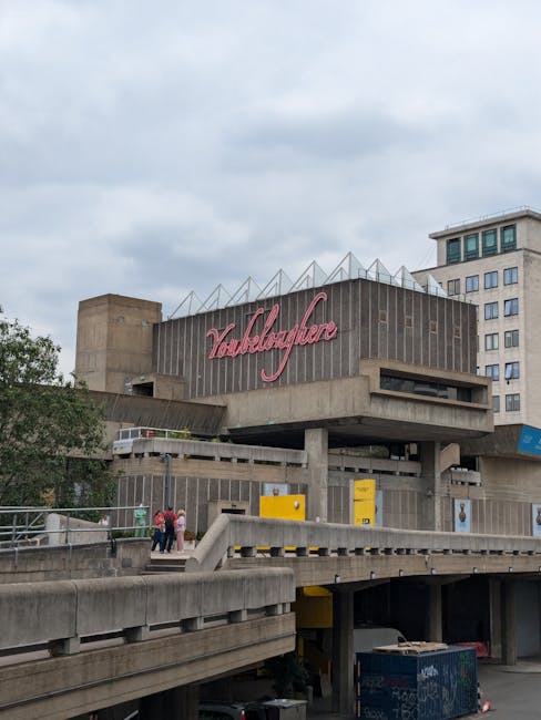 Exterior view of the Vallebierme building in Kingston upon Thames, featuring a concrete structure with multiple levels, large windows, and a prominent pink neon sign displaying the name. The surrounding area includes a walkway with a few pedestrians, a railing, and a parking area underneath. The sky is overcast, casting diffuse light over the scene, with no visible cleaning equipment or surfaces in the image. This image showcases the building’s architectural design and urban setting, relevant for the context of commercial or building maintenance. For surface cleaning or deep cleaning services in Kingston upon Thames, clients can rely on [COMPANY_NAME].