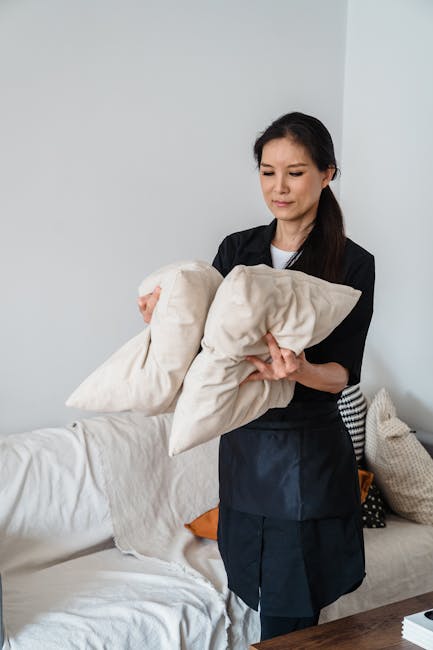 A woman with dark hair tied back, dressed in a black uniform, is holding two beige cushion covers or pillowcases and is in the process of cleaning or preparing textiles in a bright, modern living room. The room features a white sofa with a beige and a black-and-white patterned cushion, and a light-colored wall with soft natural lighting. The scene depicts domestic surface and fabric cleaning, emphasizing hygiene and maintenance, and is associated with professional cleaning services from Cleaners Kingston upon Thames as mentioned on the page about upholstery cleaning in Kingston upon Thames.