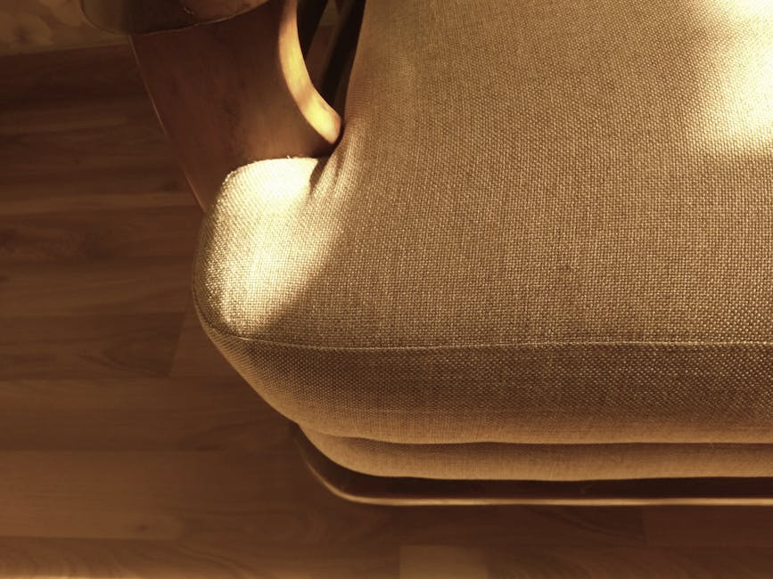Close-up of a beige fabric-upholstered armchair with a textured surface, situated on a wooden floor in a well-lit room. The chair's soft, clean cushions show no visible dust or stains, indicating recent surface cleaning or deep cleaning conducted by Cleaners Kingston upon Thames. Part of a dark wooden side table is visible on the left side, highlighting the arrangement of furniture in a domestic setting. The lighting accentuates the smooth, polished finish of the upholstery, emphasizing the hygiene and maintenance of the seating area as part of professional cleaning services visible on the page for Canbury Gardens upholstery cleaning Kingston upon Thames.